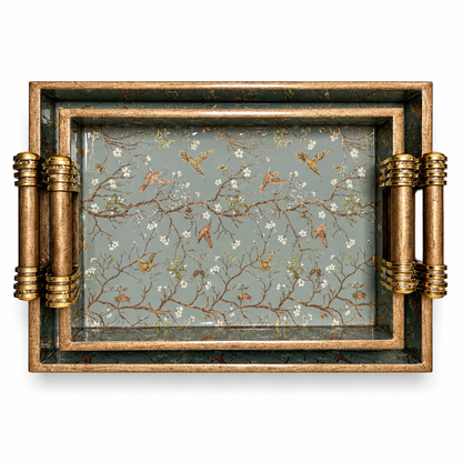 Top view of two nested wooden serving trays with sage green bird and floral patterns and gold-accented handles on a white background.